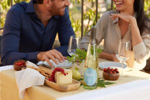 CantineMaschio_pranzo_vino: Due persone pranzano a un tavolo all’esterno bevendo una bottiglia di vino bianco frizzante di Cantine Maschio.
Tavola_apparecchiata_vino: Una tavola apparecchiata con una bottiglia di vino bianco frizzante di Cantine Maschio.